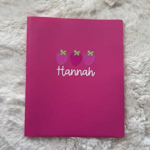 May include: A bright pink binder with the name "Hannah" in white script. Above the name are three embroidered strawberries in pink and green. The binder is closed and sits on a fluffy white surface.