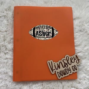May include: An orange folder featuring a central football-shaped decal with a camouflage pattern and the name Asher in black text. A wooden cutout piece is attached to the bottom right corner with the text Kinsley DOWDY CO in black script.