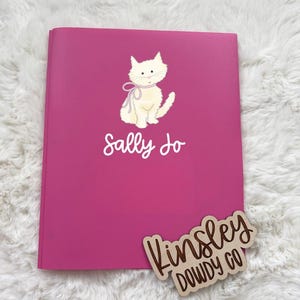 May include: A bright pink notebook with the name "Sally Jo" in white script. A cream-colored cat illustration with a pink bow is above the name. A wooden tag with the text "Kinsley Dowdy Co" is in the lower right corner.