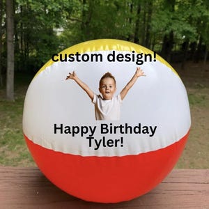 May include: A large, inflatable beach ball with a custom design. The ball is primarily white, with yellow and red bands. The text "custom design!" and "Happy Birthday Tyler!" are printed on the ball. A young child with arms raised is in the center.