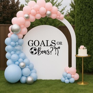 May include: A white archway decorated with pink, silver, and blue balloons, with the text "GOALS or Bows?" A small cake sits on a gold stand. The scene is set on a green lawn, suggesting a gender reveal party or baby shower.