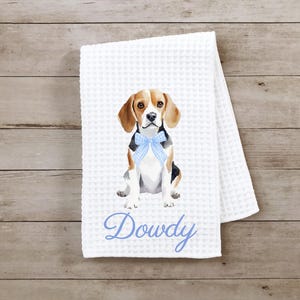 May include: White waffle weave kitchen towel with a watercolor illustration of a beagle dog wearing a blue bow. The towel is personalized with the name "Dowdy" in blue embroidery.
