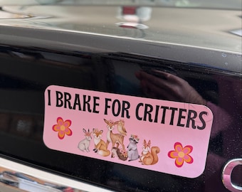 Op de afbeelding: Een roze autobumper sticker met de tekst "I Brake For Critters" en een rij cartoon bosdieren, waaronder een konijn, een vos, een hert, een uil, een wasbeer, een egel en een eekhoorn. De sticker heeft twee roze bloemen met gele centra aan weerszijden van de dieren.