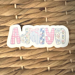 May include: A die-cut sticker featuring the name Adalyn in a playful font. Each letter displays a different pattern, including pink gingham, blue and white stripes, and floral prints in shades of blue, pink, and green, with a small pink bow.