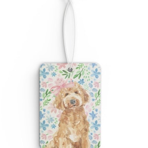 May include: A car air freshener featuring a watercolor illustration of a golden doodle dog with a pink bow, surrounded by a floral pattern of pink, blue, and green flowers.