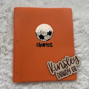 May include: An orange binder featuring a central graphic of a soccer ball with a leopard print pattern and white icing-like drips on top. The name Charles is printed in black below the ball. A wooden sign reads Kinsley DOWDY CO in script.