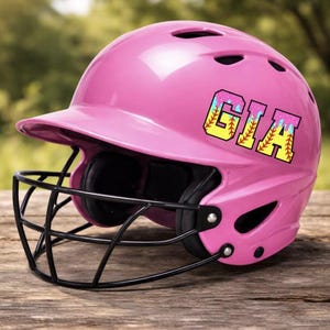 May include: A pink softball helmet with a black faceguard. The helmet has the name "GIA" in a colorful, baseball-themed font. The helmet is sitting on a wooden surface, with a blurred green background.