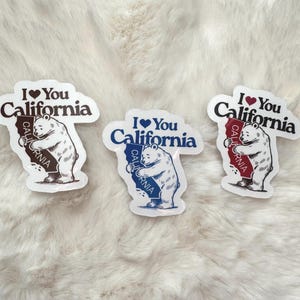 May include: Three white stickers with a brown bear hugging a California outline. Each sticker says "I ♥ You California" with a red heart.