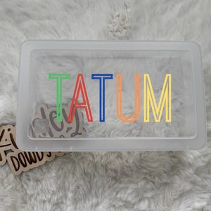 May include: A clear rectangular container with the name "TATUM" in colorful letters. The letters are green, red, blue, orange, and yellow. A wooden cutout with the text "Ki dowd" is visible.