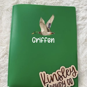 May include: A green notebook with the name "Griffen" in white text and a wooden cutout that says "Kinsley Dowdy Co". The notebook features a watercolor illustration of a flying duck.