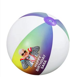 May include: A white beach ball with a rainbow gradient design. The ball has a picture of a woman wearing sunglasses and a striped shirt. The text "HAPPY BIRTHDAY SARA" is printed on the ball.