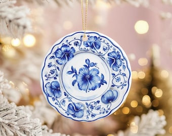 May include: A round, white ceramic ornament with a blue floral design. The ornament features a central flower and surrounding floral patterns. It hangs from a gold string, perfect for holiday decor. The ornament is approximately 4 inches in diameter.