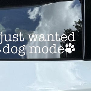 May include: White vinyl car decal with the text "I just wanted dog mode" and paw print graphics. The decal is applied to a car window, with a reflection of clouds and trees visible.
