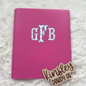 May include: A hot pink notebook with a monogram in a floral pattern. The monogram features the letters "GFB". A wooden tag with the text "Kinsley Dowdy Co" is in the lower right corner.