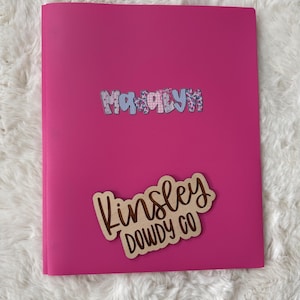 May include: A bright pink folder with two decorative name tags. The top tag says "MaKaylyn" in a patterned font. The bottom tag is wooden and reads "Kinsley Dowdy Co" in a cursive font.