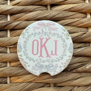 May include: A round white car coaster with a floral wreath design. The coaster features the letters OKI in pink, surrounded by a wreath of blue and pink flowers. Pink and blue bows are at the top and bottom.