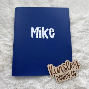 May include: A dark blue rectangular book with the name Mike printed in a light blue and white checkered font on the front cover. A small wooden sign with the text Kinsley Dowdy Co in a brown script font is positioned at the bottom right.