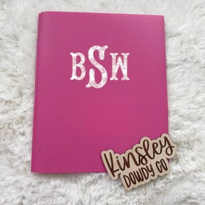 May include: A bright pink notebook with the initials "BSW" in a glittery font. A wooden tag with the text "Kinsley DOWDY CO" is placed on the bottom right corner. The notebook is on a white, fluffy surface.