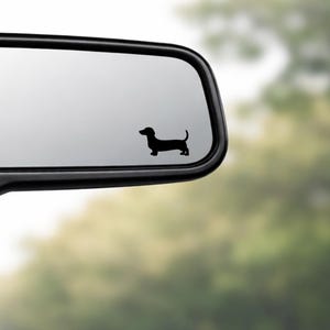 May include: A black dachshund silhouette decal on a car's rearview mirror. The dog is facing right, with a long body and short legs. The mirror is black, and the background is a blurred view of green trees.