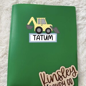 May include: A green notebook with a yellow and white checkered excavator graphic and the name "TATUM" on the front. A wooden sign with the name "Kinsley DOWDY CO" is placed on the bottom right corner.