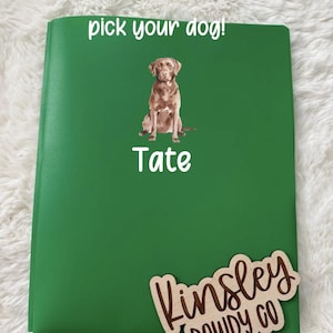 May include: A green notebook with the text "pick your dog!" and the name "Tate" below a brown dog illustration. A wooden sign with the name "Kinsley Dowdy Co" is attached to the notebook.