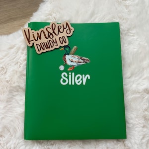 May include: A green notebook with the name "Siler" in white lettering and a duck illustration. A wooden tag with the text "Kinsley Dowdy Co" is placed on top of the notebook, resting on a white, fluffy surface.