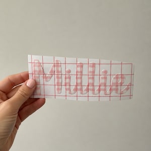 Könnte beinhalten: Ein individueller Vinyl-Aufkleber mit dem Namen Millie in einem rosa-weißen Vichy-Karomuster. Der Schriftzug befindet sich auf einer rechteckigen weißen Trägerfolie mit rotem Raster, die für die Verwendung als personalisiertes Etikett gedacht ist.