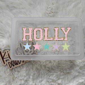 May include: A clear plastic storage box with the name "HOLLY" in pink glitter letters outlined in gold. Below the name are four pastel-colored stars. A wooden cutout with text is visible on the left.