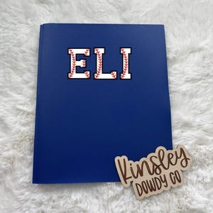 May include: A navy blue notebook with the name "ELI" in a baseball-themed font. A wooden name tag with the name "Kinsley DOWDY CO" is also visible.