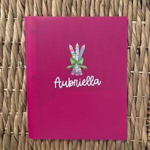 May include: A bright pink plastic folder featuring a central graphic of three pencils tied together with a green bow. Below the pencils, the name Aubriella is printed in white script lettering. The folder has two visible holes on the left side.