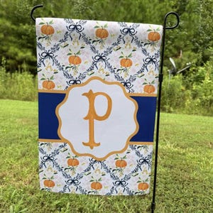 May include: A rectangular garden flag featuring a repeating pattern of orange pumpkins, dark blue bows, and delicate floral vines on a white background. A horizontal dark blue band crosses the center with a gold-outlined white frame containing a gold letter P.