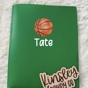 May include: Green notebook with a basketball graphic and the name "Tate" in white. A wooden tag with the name "Kinsley Dowdy Co" is attached to the notebook.