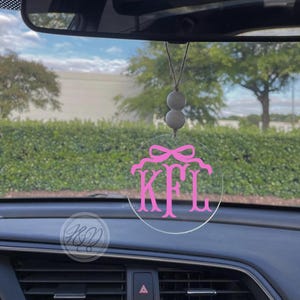 May include: A pink and white car charm with the monogram "KFL" in a circle. The charm is hanging from the rearview mirror of a car.