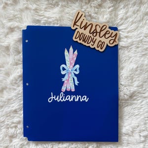 May include: A royal blue binder with a pencil illustration and the name "Julianna" in white script. A wooden name tag that says "Kinsley Dowdy Co" is on the top right.