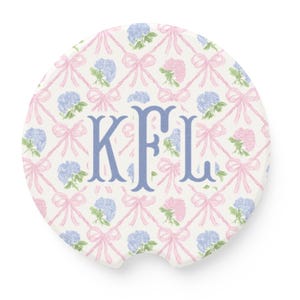 May include: Round car coaster with a white background, featuring a pattern of pink bows and blue and pink hydrangeas. The center displays the blue monogram letters KFL.