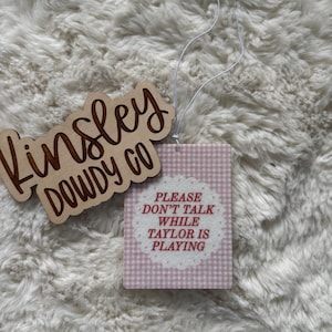 May include: Wooden sign with the text "Kinsley Dowdy Co" in a cursive font. A pink and white gingham air freshener with the text "Please don't talk while Taylor is playing" is attached with a white string.