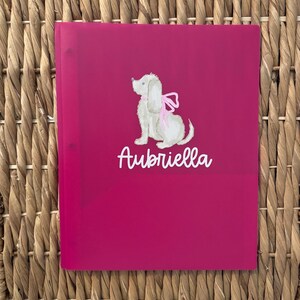 May include: A bright magenta plastic folder featuring a white graphic of a seated dog with a pink bow around its neck. The name Aubriella is printed in white cursive script below the dog illustration on the front cover of the folder.