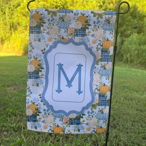 May include: A rectangular garden flag featuring a repeating pattern of orange and light blue pumpkins, white flowers, and blue plaid squares. A central light blue scalloped frame contains a large, bold, blue capital letter M in the center.