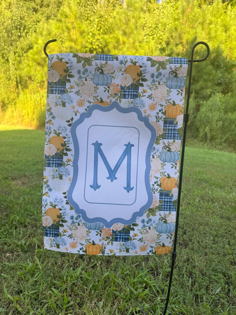 K&ouml;nnte beinhalten: Eine rechteckige Gartenflagge mit einem wiederkehrenden Muster aus orangefarbenen und hellblauen K&uuml;rbissen, wei&szlig;en Blumen und blauen Karomustern. Ein zentraler, hellblauer, gewellter Rahmen enth&auml;lt einen gro&szlig;en, blauen Gro&szlig;buchstaben M.