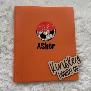 May include: An orange three-ring binder featuring a graphic of a soccer ball with a red, sprinkle-covered top half. The name Asher is printed in black below the graphic. A wooden cutout with the text Kinsley DOWDY CO is placed on the binder.