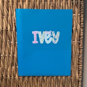 May include: A bright blue notebook featuring the name IVEY centered on the front cover. The letters are decorated with various patterns including pink gingham, blue and white stripes, and floral prints, with a small pink bow on the letter V.