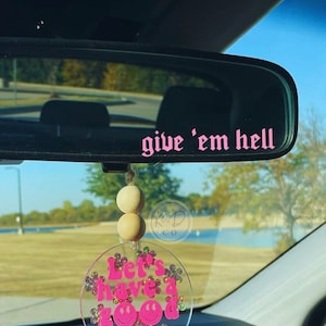 May include: Pink and white car charm with the text "Let's have a good day" and a smiley face. The charm is hanging from a rearview mirror with a pink sticker that says "give 'em hell".