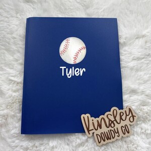 May include: A navy blue notebook with a baseball graphic and the name "Tyler" in white script. A wooden sign with the text "Kinsley Dowdy Co" is also present.