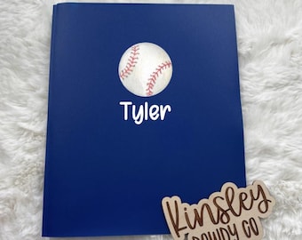 以下が含まれることがあります： 野球のグラフィックと白いスクリプトで「Tyler」という名前が入った濃い青色のノート。 「Kinsley Dowdy Co」というテキストが入った木製の看板も表示されています。