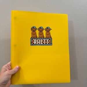 May include: A bright yellow binder with a graphic of three golden retriever puppies wearing blue caps. The puppies are above a sign that reads "RHETT" in white letters. The binder has three holes and is held by a hand.