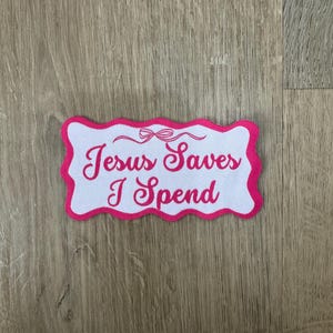 May include: A white rectangular patch with a pink scalloped border. The patch features the embroidered text "Jesus Saves I Spend" in pink script. A small bow design is above the text. The patch is likely for clothing or accessories.