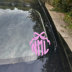 May include: Pink monogram decal with a bow on a black car window. The monogram reads "NML".