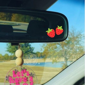 May include: A pink and white car charm with the text "Let's Have a Good Day" and two smiley faces. The charm is hanging from a rearview mirror with two wooden beads. Two red strawberries are visible in the rearview mirror.