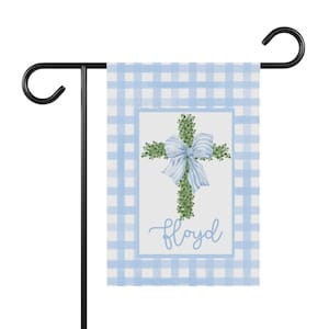May include: A light blue and white gingham garden flag with a cross made of green leaves and a light blue bow. The word "Floyd" is written in cursive script. The flag is hanging on a black metal stand.