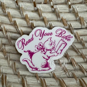 May include: A white sticker with a pink outline and a pink illustration of a cat reading a book. The text "Read Your Bible" is written in pink cursive.